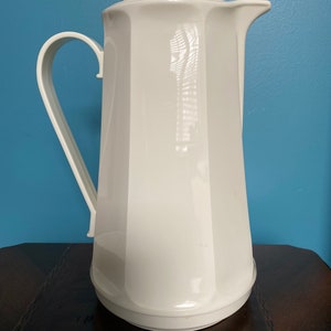 May include: A white, vintage thermal jug with a tall, faceted body and a curved handle. The jug has a spout for pouring and a lid with a decorative top. It is likely made of plastic or a similar material.