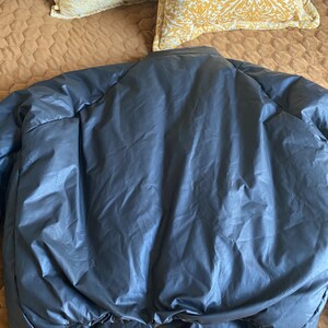 May include: A dark gray puffer jacket, viewed from the back, lies on a bed. The jacket has a smooth, shiny texture and a rounded collar. The bottom hem is elasticized. Two decorative pillows with a yellow and white pattern are in the background.