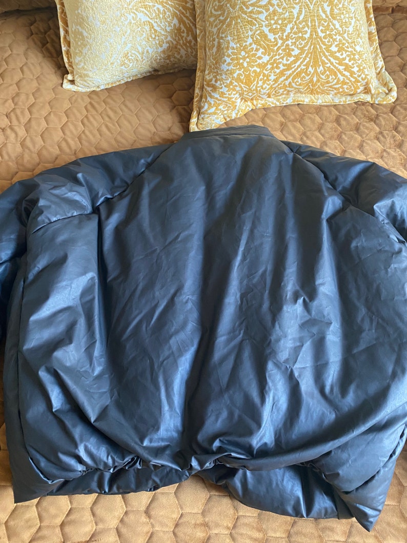 May include: A dark gray puffer jacket, viewed from the back, lies on a bed. The jacket has a smooth, shiny texture and a rounded collar. The bottom hem is elasticized. Two decorative pillows with a yellow and white pattern are in the background.