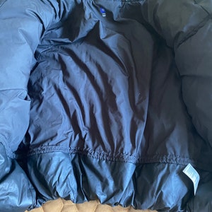 May include: A dark navy blue puffer jacket, viewed from the inside. The jacket has a quilted design and a drawstring at the waist. A small tag is visible near the bottom hem. The jacket is made of a shiny, smooth material.