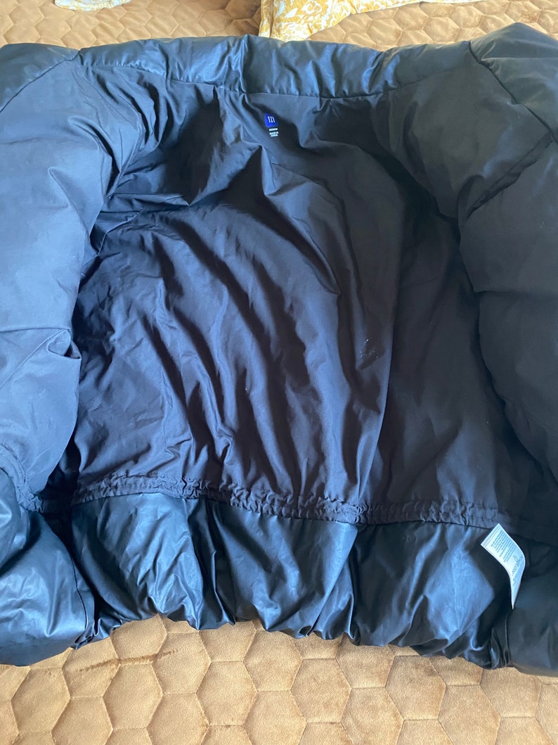 May include: A dark navy blue puffer jacket, viewed from the inside. The jacket has a quilted design and a drawstring at the waist. A small tag is visible near the bottom hem. The jacket is made of a shiny, smooth material.