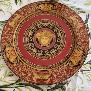 Puede incluir: Un plato decorativo con un centro rojo y un dise&ntilde;o de cabeza de Medusa dorada. El plato presenta adornos dorados y negros y un borde dorado. El borde exterior es rojo con detalles dorados.