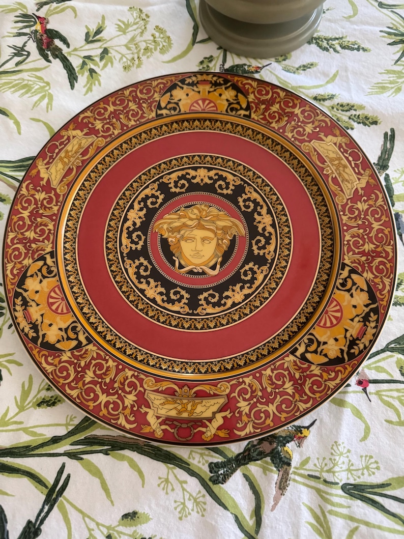 Puede incluir: Un plato decorativo con un centro rojo y un dise&ntilde;o de cabeza de Medusa dorada. El plato presenta adornos dorados y negros y un borde dorado. El borde exterior es rojo con detalles dorados.
