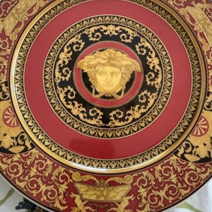 Puede incluir: Un plato decorativo con un dise&ntilde;o rojo, negro y dorado. El plato presenta un medall&oacute;n central con una cara dorada, rodeado de intrincados patrones y un borde dorado. El borde exterior tiene un dise&ntilde;o de desplazamiento repetido.