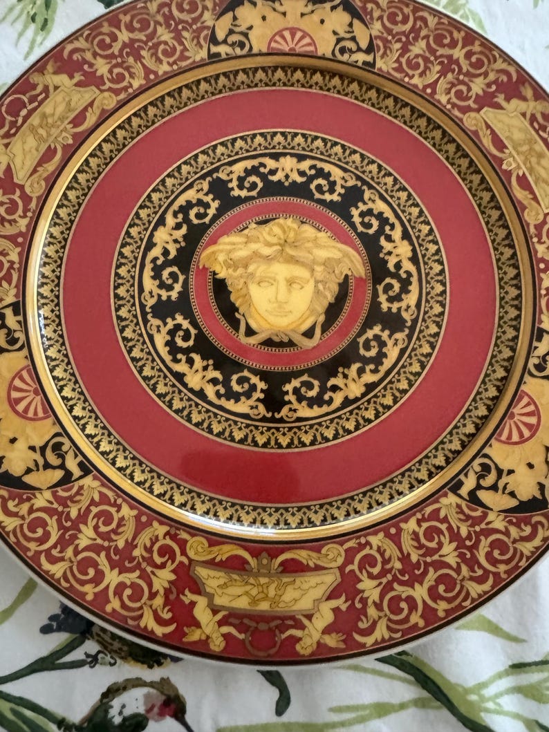 Puede incluir: Un plato decorativo con un dise&ntilde;o rojo, negro y dorado. El plato presenta un medall&oacute;n central con una cara dorada, rodeado de intrincados patrones y un borde dorado. El borde exterior tiene un dise&ntilde;o de desplazamiento repetido.