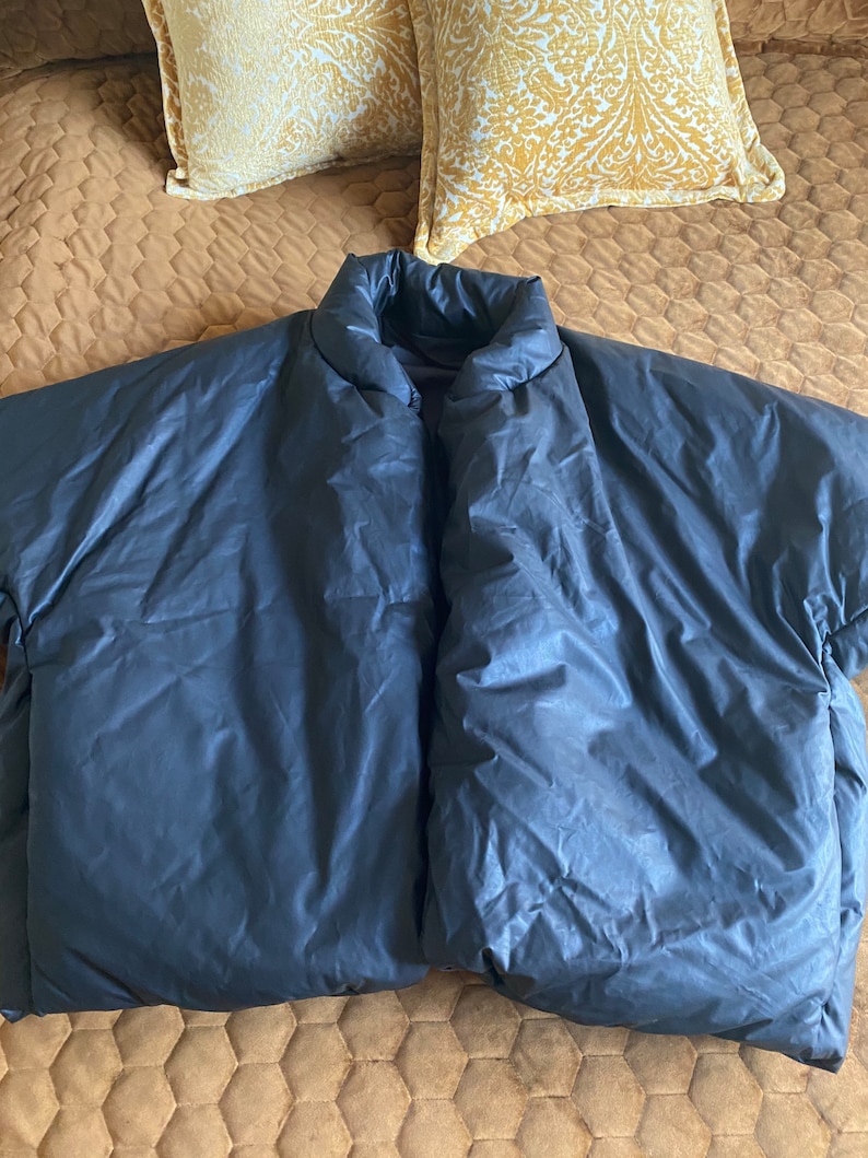 May include: A dark navy blue puffer jacket with a high collar, spread out on a bed. The jacket has a shiny, smooth texture. Two decorative yellow pillows with a floral pattern are in the background.