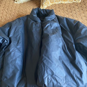 May include: A dark blue puffer jacket with a high collar. The jacket is made of a shiny, quilted material. The jacket is lying on a brown bedspread with two yellow pillows in the background.