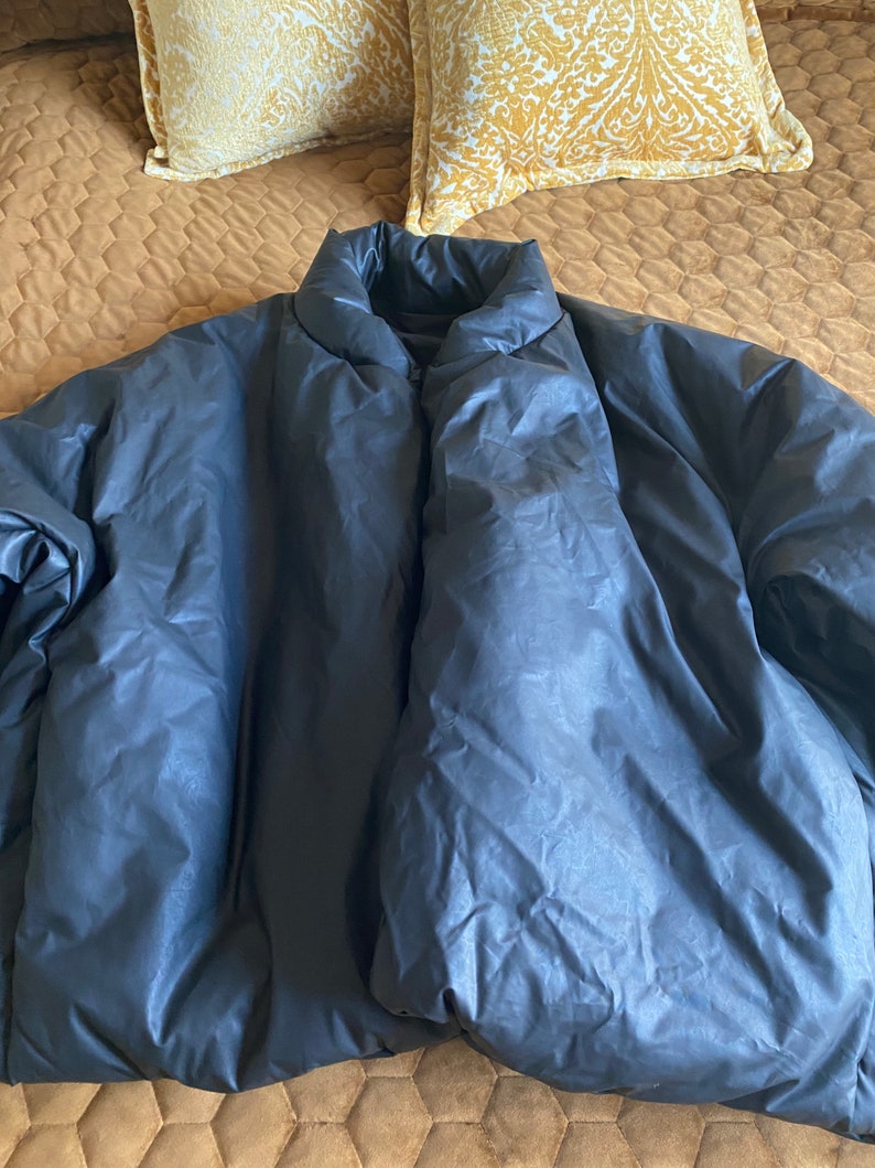 May include: A dark blue puffer jacket with a high collar. The jacket is made of a shiny, quilted material. The jacket is lying on a brown bedspread with two yellow pillows in the background.