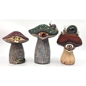 May include: Three sculpted mushroom figurines with textured stems and caps. The left mushroom has a maroon cap and yellow eyes. The center mushroom is green with a single orange eye and a snail. The right mushroom is brown with a single eye and a snail.