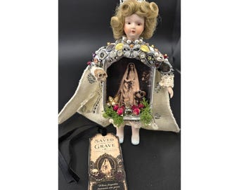 May include: A porcelain doll with blonde curls wearing a silver crown and a cape adorned with various metallic trinkets and jewels. The doll holds a framed image of a person in a veil, accented with small dried red roses and green moss. A black tag reads SAVED FROM THE GRAVE.