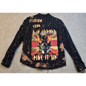 May include: A black and grey plaid long-sleeve button-down shirt with a bleached effect. The back features a large rectangular patch with the text THE STADIUM TOUR DEF LEPPARD FIRE IT UP and a graphic of a skull with flames in the eye sockets.