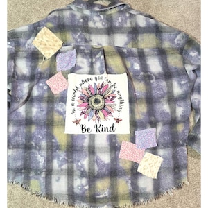 May include: A long-sleeve button-down shirt with a purple, gray, and white plaid pattern featuring a bleached effect. A central white patch displays a colorful sunflower graphic with the text In a world where you can be anything Be Kind.