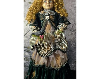 May include: A doll with curly blonde hair and pale skin wearing a dark green velvet jacket over a dress with a striped bodice and a long, layered skirt. The doll holds a small black bird and a cluster of tiny red berries in its hands.