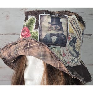 May include: A brown bucket hat featuring frayed edges and a patchwork design. The hat displays a central image of a cat wearing a top hat and tuxedo, a red rose, a bird illustration, and a label with the text YOU ARE ENOUGH. The underside is plaid.