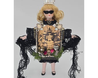 May include: A porcelain doll with blonde hair wearing a silver crown and a black eye mask. The doll features a central rectangular frame displaying a sepia-toned image of three figures, accented with green moss, pink flowers, a skull, and black lace.
