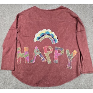 May include: A long-sleeve, mineral-washed maroon shirt featuring a decorative, multi-colored fabric rainbow applique above the word HAPPY. Each letter of the word is crafted from a different floral and patterned fabric patch sewn onto the shirt.