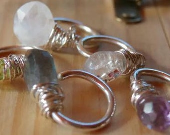 May include: A collection of silver wire-wrapped rings, each featuring a single faceted gemstone. The stones include clear quartz, light green peridot, dark grey labradorite, and purple amethyst, all secured with silver wire coils.
