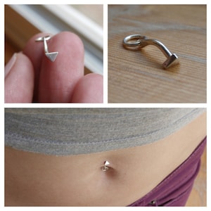 Könnte beinhalten: Ein silberner Bauchnabelring mit einem Dreieck und einer gebogenen Stange. Der Ring ist auf dem Bauchnabel einer Person zu sehen.