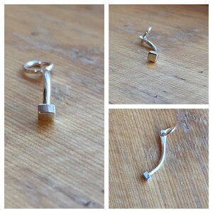 May include: A silver charm with a curved shape and a square at the end. The charm has a loop at the top for attaching to a bracelet or necklace.