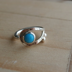 May include: A silver ring with a turquoise stone set in a unique, abstract design. The ring has a textured finish and a delicate, openwork design.