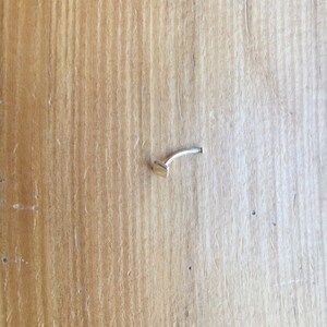 May include: A small, silver nose ring with a square-shaped stud. The ring is on a wooden surface.
