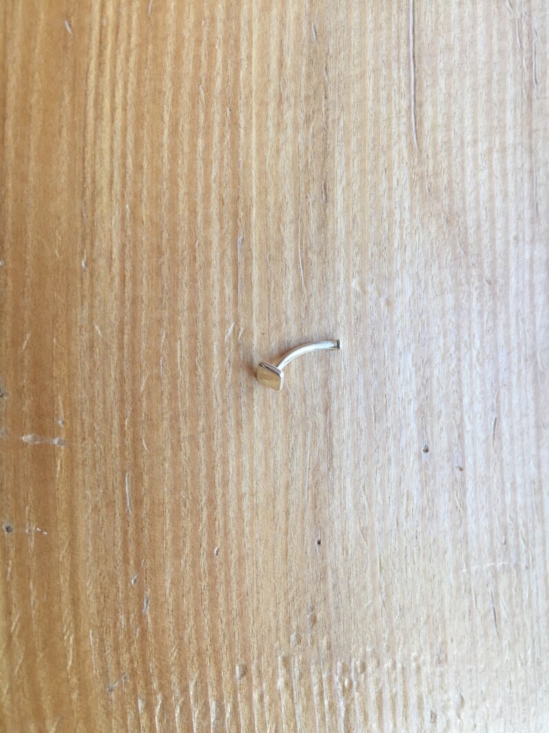 May include: A small, silver nose ring with a square-shaped stud. The ring is on a wooden surface.