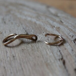 May include: Two gold-toned metal clasps, one with a curved shape and the other a simple ring, are shown on a light brown wooden surface.