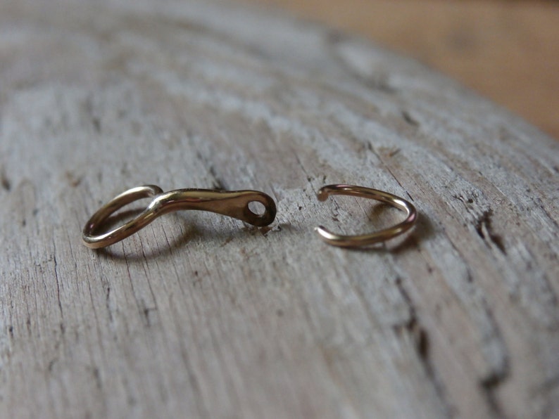 May include: Two gold-toned metal clasps, one with a curved shape and the other a simple ring, are shown on a light brown wooden surface.