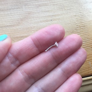 May include: A silver nose ring with a square-shaped stud. The ring is curved and has a small, flat end.
