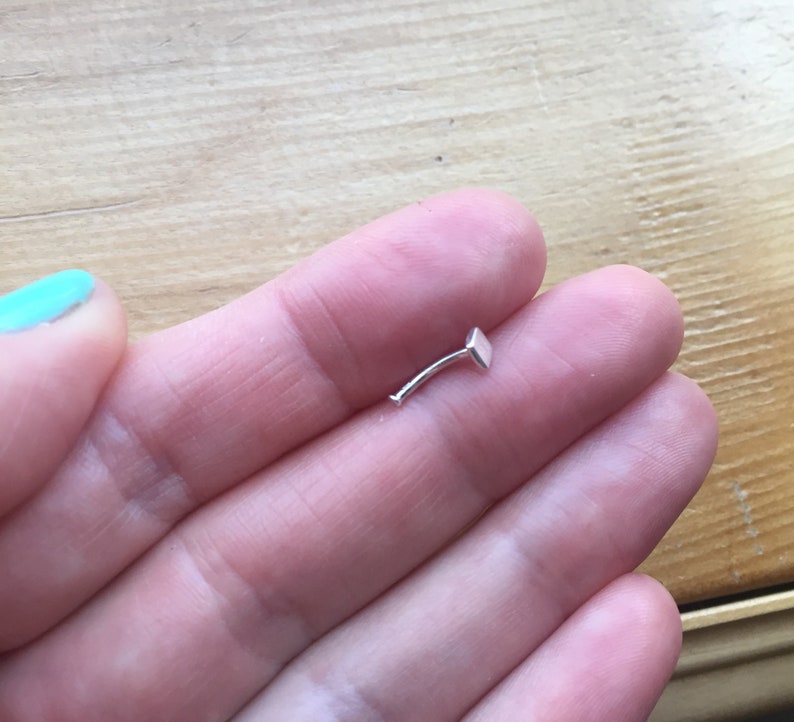 May include: A silver nose ring with a square-shaped stud. The ring is curved and has a small, flat end.