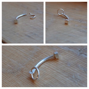 May include: A silver ear cuff with a square-shaped end. The cuff is bent into a curved shape and has a small ring at the end.