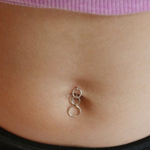 Puede incluir: Un piercing de ombligo plateado con tres círculos conectados que cuelgan.