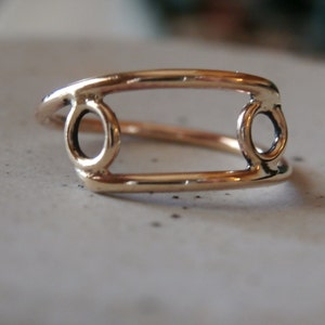 May include: A gold-colored ring with a unique design. The ring features a rectangular shape with two circular accents on each end. The ring is made of a shiny, metallic material and is set against a neutral background.