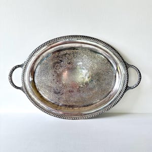 May include: Oval silver serving tray with ornate floral detailing and rope-style edging. The tray has two loop handles and a reflective surface. A vintage piece for serving food or displaying items.