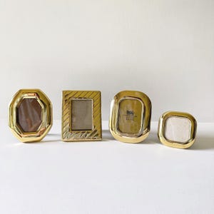 May include: Four polished gold-tone metal picture frames of varying shapes including an octagon, a rectangle with diagonal etched lines, an oval, and a square with rounded corners. One frame contains a label reading Solid Brass 2.5 x 3.5 inches.
