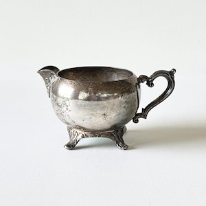 May include: Antique silver creamer with a spout and ornate handle. The creamer sits on three decorative feet. The silver has a tarnished appearance, adding to its vintage charm. A classic piece for serving cream or milk.