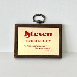 May include: A framed plaque with the name "Steven" in red script. The text "HIGHEST QUALITY" is below the name, with a quote from Psalm 8:5. The frame is brown, and it has a decorative handle at the top.