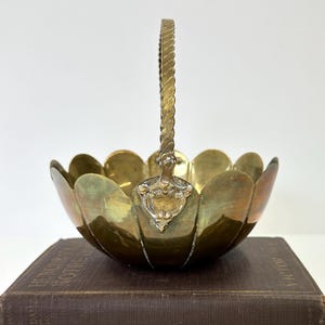 May include: A small, brass, scalloped bowl with a twisted handle. The bowl has a decorative design on the front.