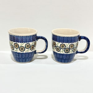 May include: Two ceramic mugs with a blue polka dot pattern and a band of floral designs in green, yellow, and brown. The mugs have a cream-colored interior and a curved handle. The mugs are on a white surface.