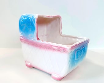 May include: A ceramic baby cradle planter in white, blue, and pink. The cradle features a diamond pattern on the sides, a scalloped pink trim, and blue accents. The planter has a vintage aesthetic.