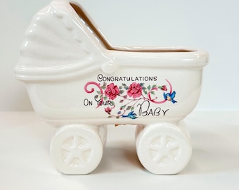May include: A white ceramic baby carriage planter with a decorative floral design and the words "Congratulations On Your Baby." The carriage has four wheels and a hood. The floral design includes pink roses and blue birds.