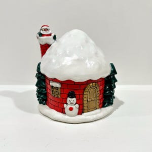 May include: A ceramic Christmas house figurine with a red brick exterior, a snow-covered roof, and a chimney with Santa Claus. The house features a snowman, a gold door, and green Christmas trees.