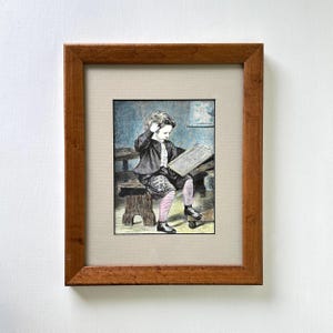 May include: Framed vintage print of a young boy seated on a bench, engrossed in reading a slate. The artwork features a brown wooden frame, a beige mat, and a detailed illustration in shades of gray, black, and pink. The boy is wearing a jacket, patterned shorts, and striped socks.