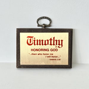 May include: A framed plaque with the name "Timothy" in red script. The text "HONORING GOD" and a Bible verse are below the name. The frame is dark brown, and the plaque has a decorative metal handle.