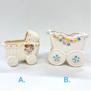 May include: Two white ceramic baby carriages, one shaped like a bassinet and the other a traditional carriage, both with colourful floral accents and wheels. The carriages are perfect for holding small plants or other decorative items.