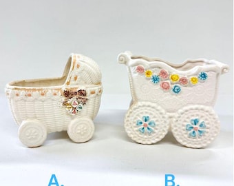 May include: Two white ceramic baby carriages, one shaped like a bassinet and the other a traditional carriage, both with colorful floral accents and wheels.  The carriages are perfect for holding small plants or other decorative items.