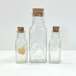 May include: Three clear glass bottles with cork stoppers. The bottles are rectangular with embossed designs and text. The largest bottle reads "POUND PURE HONEY". The other bottles have text that reads "Grapes Pure" and "Ollies Pure".