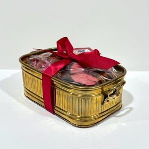 May include: A rectangular brass planter with vertical ribbed sides and two side handles. The container is filled with dried pink and brown potpourri sealed in a clear plastic bag and is wrapped with a wide, bright red satin ribbon tied in a bow.