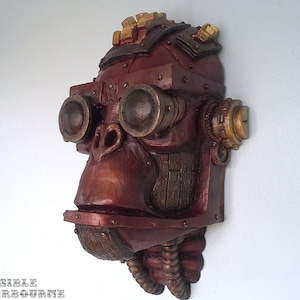 May include: A wall-mounted sculpture of a primate head in a metallic red finish with gold accents. The figure wears large circular goggles over the eyes, features mechanical gear-like details on the sides, and has textured tubing extending from the chin.