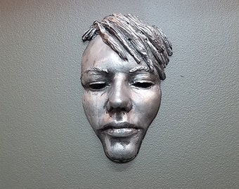 May include: A silver-colored metal sculpture of a face with a textured hairstyle. The face has a neutral expression with closed eyes and a slightly parted mouth.